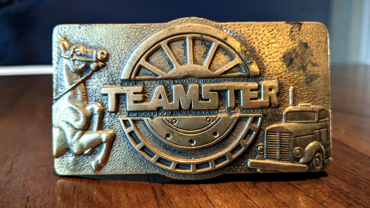 Close-up of a vintage Teamsters Union belt buckle featuring raised motifs of a horse-drawn carriage and a classic semi-truck within a circular design, symbolizing the transportation and labor history of the organization