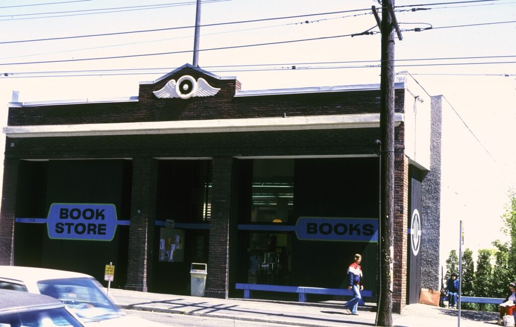 The Seattle Central Community College bookstore, circa 1978, located in the historic building of the former Teamsters Union headquarters on Broadway, Capitol Hill. The facade features bold 'BOOK STORE' and 'BOOKS' signage on a vintage brick structure with large windows, with a pedestrian passing by and a blue car parked in front.